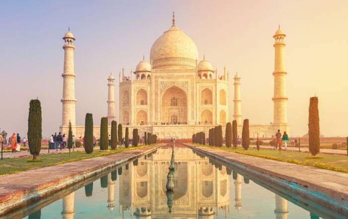 Mausoleul Taj Mahal din marmură albă se înalță maiestuos, reflectându-se într-un bazin lung de apă mărginit de copaci verzi și turiști, sub lumina caldă a răsăritului.