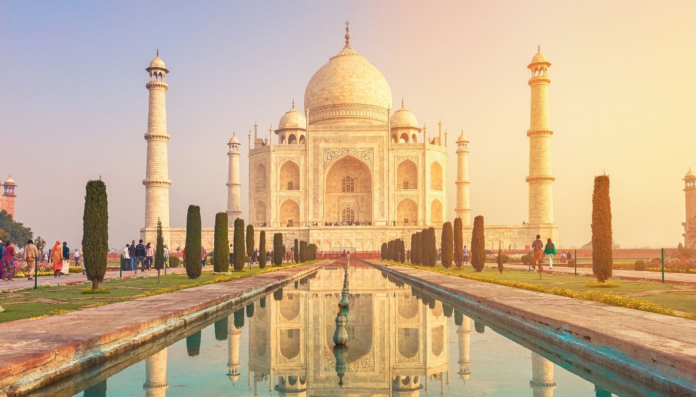 Mausoleul Taj Mahal din marmură albă se înalță maiestuos, reflectându-se într-un bazin lung de apă mărginit de copaci verzi și turiști, sub lumina caldă a răsăritului.