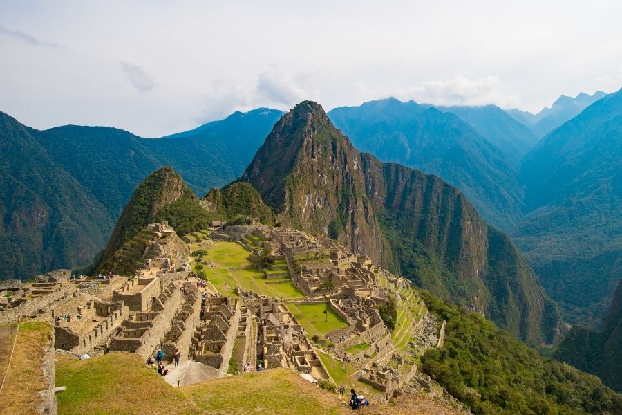 Ruinele din piatră ale cetății incașe Machu Picchu se întind pe crestele verzi, având în fundal muntele Huayna Picchu, înconjurate de piscuri abrupte sub un cer parțial noros.