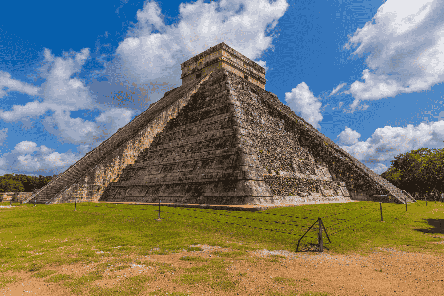 Piramida El Castillo din piatră se înalță impunător pe o pajiște verde la Chichén Itzá, sub un cer albastru presărat cu nori albi și pufoși.