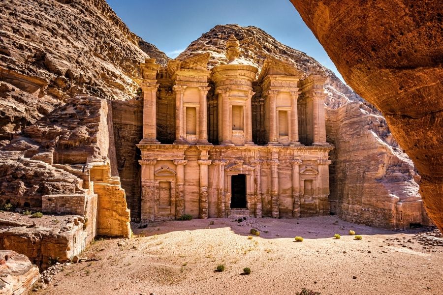 Fațada monumentului Ad-Deir (Mănăstirea) din Petra stă sculptată direct în stânca de gresie trandafirii, dominând o esplanadă aridă și însorită, flancată de munți stâncoși.