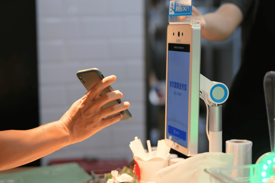 O mână ține un smartphone în fața unui terminal digital cu ecran albastru, efectuând o plată electronică într-un magazin sau restaurant modern.