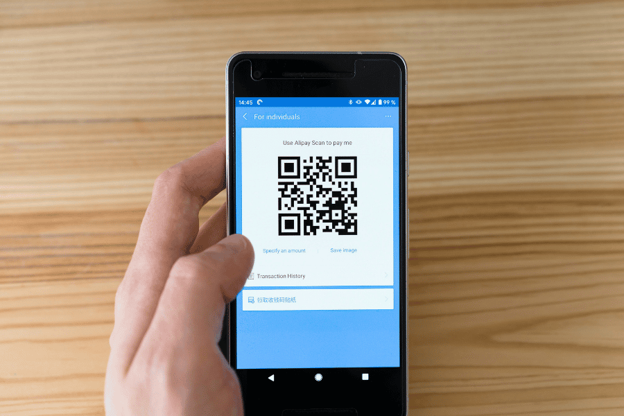 O mână ține un smartphone care afișează un cod QR de plată pe ecran, deasupra unei mese din lemn deschis la culoare.