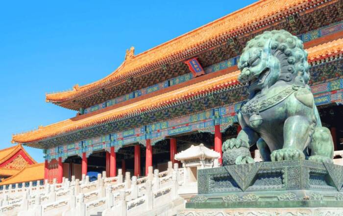 O statuie de leu din bronz păzește o clădire tradițională chinezească cu acoperiș portocaliu și stâlpi roșii, sub un cer albastru senin, în Orașul Interzis.