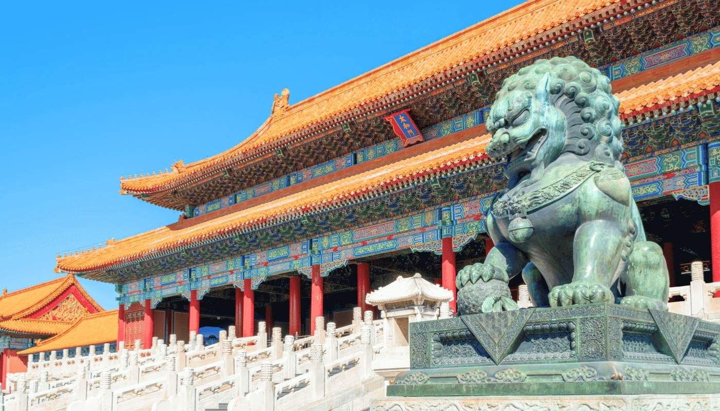 O statuie de leu din bronz păzește o clădire tradițională chinezească cu acoperiș portocaliu și stâlpi roșii, sub un cer albastru senin, în Orașul Interzis.
