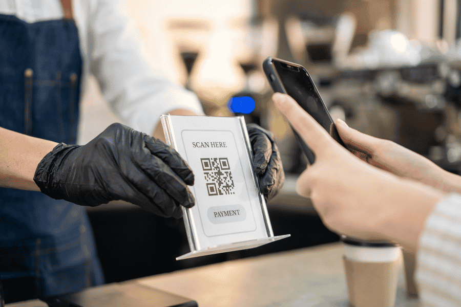 Un angajat cu mănuși prezintă un suport cu cod QR unui client care îl scanează cu smartphone-ul pentru a plăti, la tejgheaua unei cafenele.