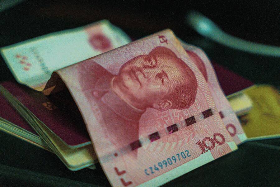 O bancnotă roz de 100 de yuani chinezești stă împăturită deasupra unui pașaport visiniu și a unor carduri bancare, pe o suprafață neagră, sugerând pregătiri de călătorie.