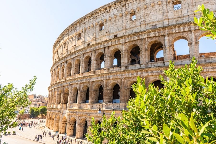 Coloseumul din Roma își înalță arcadele masive din piatră deasupra unei alei cu turiști, fiind parțial încadrat de tufișuri verzi în prim-plan, sub un cer senin.