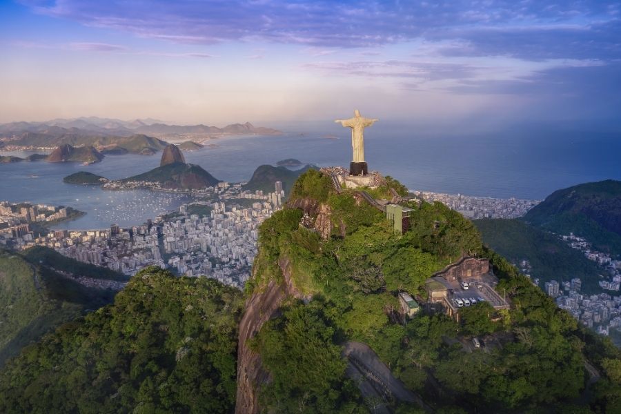 Statuia Cristos Mântuitorul veghează de pe vârful muntelui Corcovado, dominând orașul Rio de Janeiro și golful său plin de ambarcațiuni, sub un cer în nuanțe de amurg.