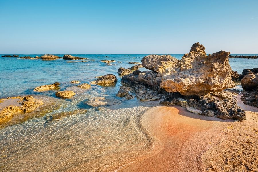 Elafonissi, Creta, Grecia. Nisipul fin cu nuanțe roz întâlnește o mare deosebit de limpede și puțin adâncă, presărată cu formațiuni stâncoase poroase pe plaja Elafonissi, sub un cer senin.