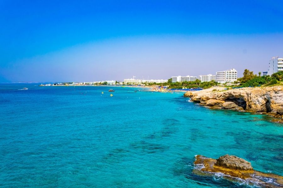Nissi Beach, Ayia Napa, Cipru. O mare turcoaz și limpede scaldă o coastă stâncoasă joasă, în timp ce hoteluri albe se întind de-a lungul plajei îndepărtate, sub un cer albastru intens în Cipru.
