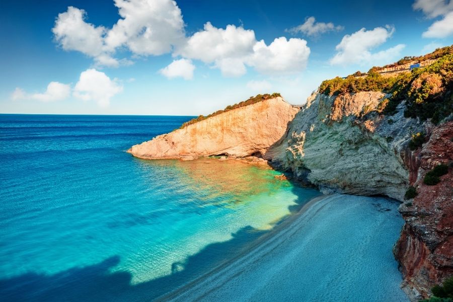 O plajă cu pietriș alb se întinde la baza unor stânci calcaroase uriașe, fiind udată de o mare turcoaz strălucitoare, sub un cer albastru cu nori pufoși. Porto Katsiki, Lefkada, Grecia