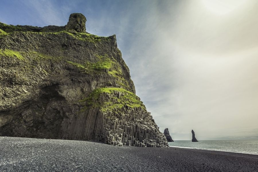 Reynisfjara, Islanda. Coloane geometrice din bazalt negru și o stâncă masivă parțial înverzită domină plaja cu nisip întunecat Reynisfjara, mărginită de mare sub un cer înnorat și dramatic.