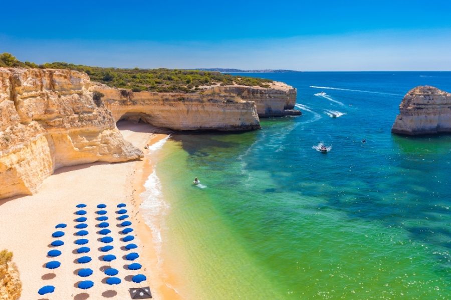 Umbrele albastre de plajă stau aliniate pe nisipul fin, flancat de stânci calcaroase și o mare turcoaz în care navighează bărci, sub un cer complet senin. Praia da Marinha, Algarve, Portugalia
