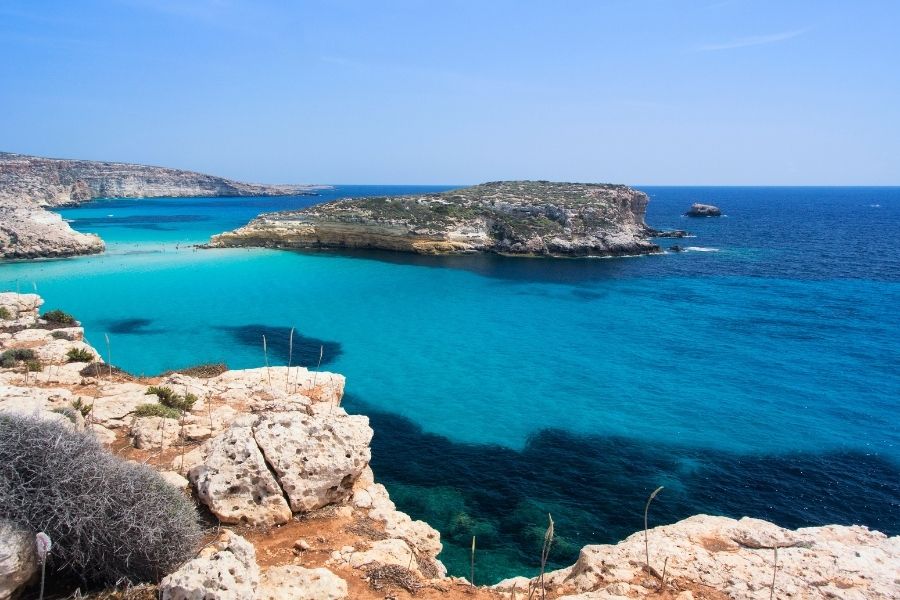 O insulă mică și stâncoasă este înconjurată de apele turcoaz și limpezi ale mării, văzută de pe coasta înaltă și aridă a insulei Lampedusa. Spiaggia dei Conigli