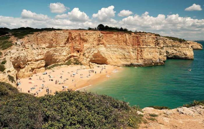 O plajă cu nisip auriu adăpostește numeroși turiști la baza unor stânci calcaroase înalte, fiind udată de o mare turcoaz, sub un cer cu nori albi. Algarve, Portugalia.
