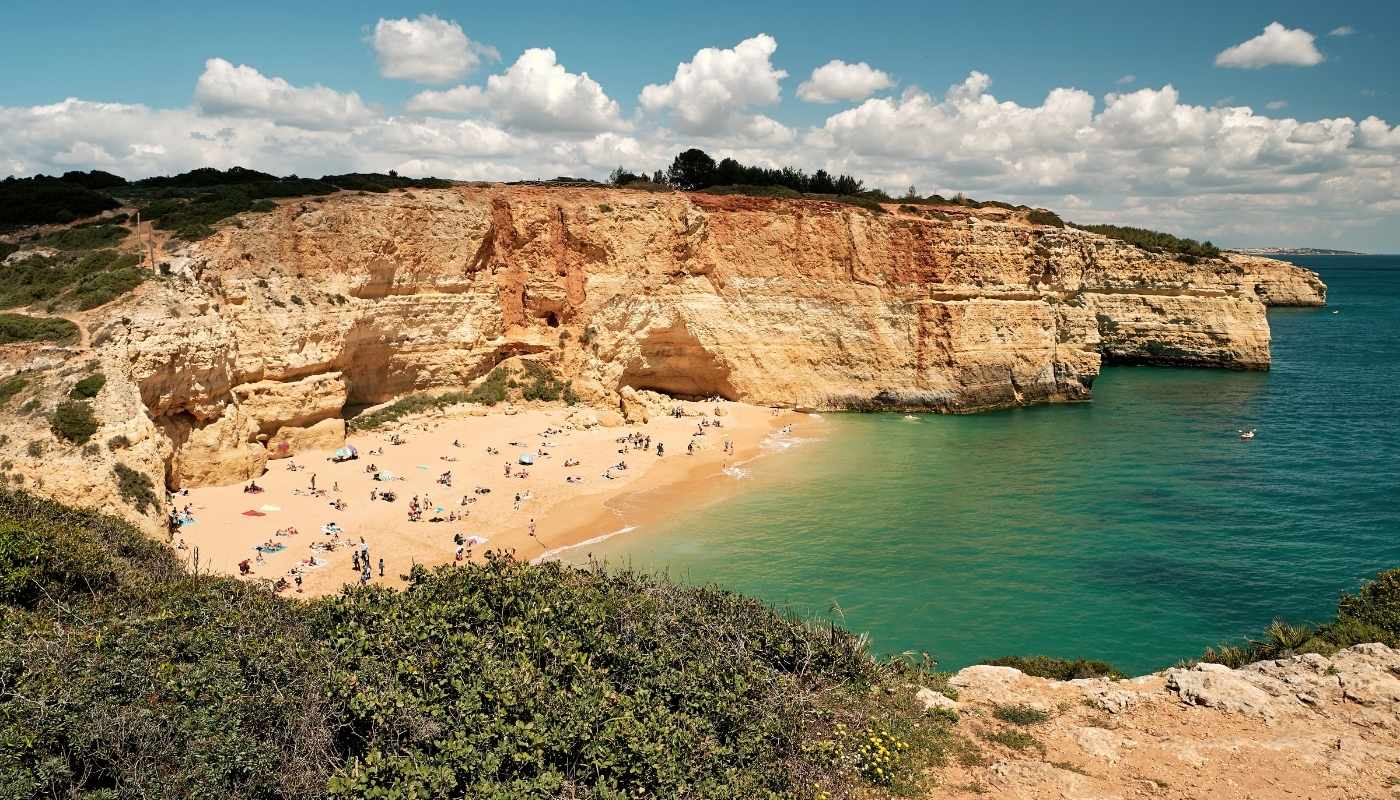 O plajă cu nisip auriu adăpostește numeroși turiști la baza unor stânci calcaroase înalte, fiind udată de o mare turcoaz, sub un cer cu nori albi. Algarve, Portugalia.