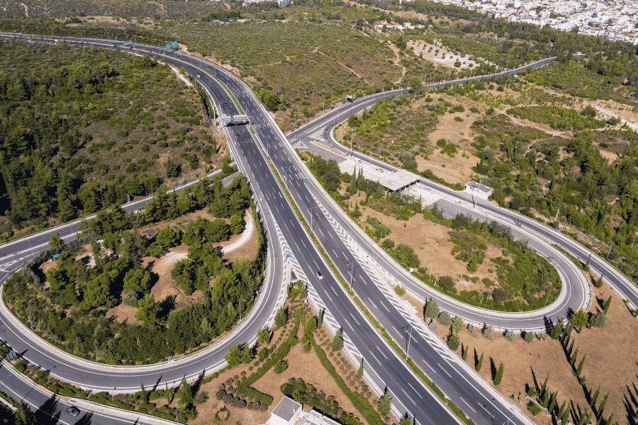 O autostradă modernă cu mai multe benzi și bucle de ieșire se șerpuiește printre dealuri verzi și vegetație, având un oraș vizibil în depărtare.