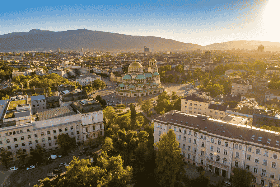 O vedere aeriană panoramică prezintă Catedrala Alexander Nevsky din Sofia, Bulgaria, situată într-o zonă urbană densă, la apus, cu munți în fundal.