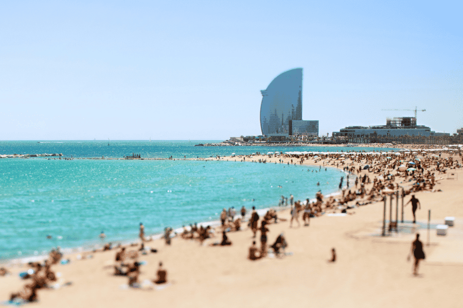 Plaja Barceloneta aglomerată de nisip se întinde de-a lungul mării turcoaz pline de oameni, având hotelul în formă de velă în fundal sub un cer senin.