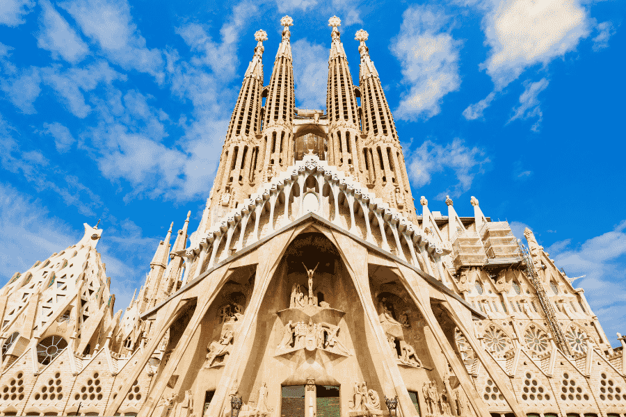 Catedrala Sagrada Família se înalță impunător cu turnurile sale sculptate, dominând cadrul sub un cer albastru cu nori razleți.