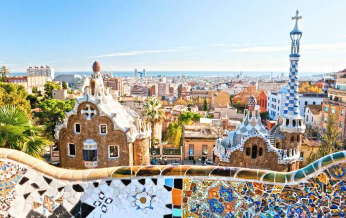 O bancă decorată cu mozaic colorat se întinde în prim-plan, oferind o priveliște asupra clădirilor din Parcul Güell și a orașului Barcelona, sub un cer senin, până la mare.