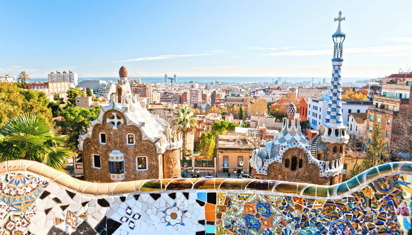 O bancă decorată cu mozaic colorat se întinde în prim-plan, oferind o priveliște asupra clădirilor din Parcul Güell și a orașului Barcelona, sub un cer senin, până la mare.