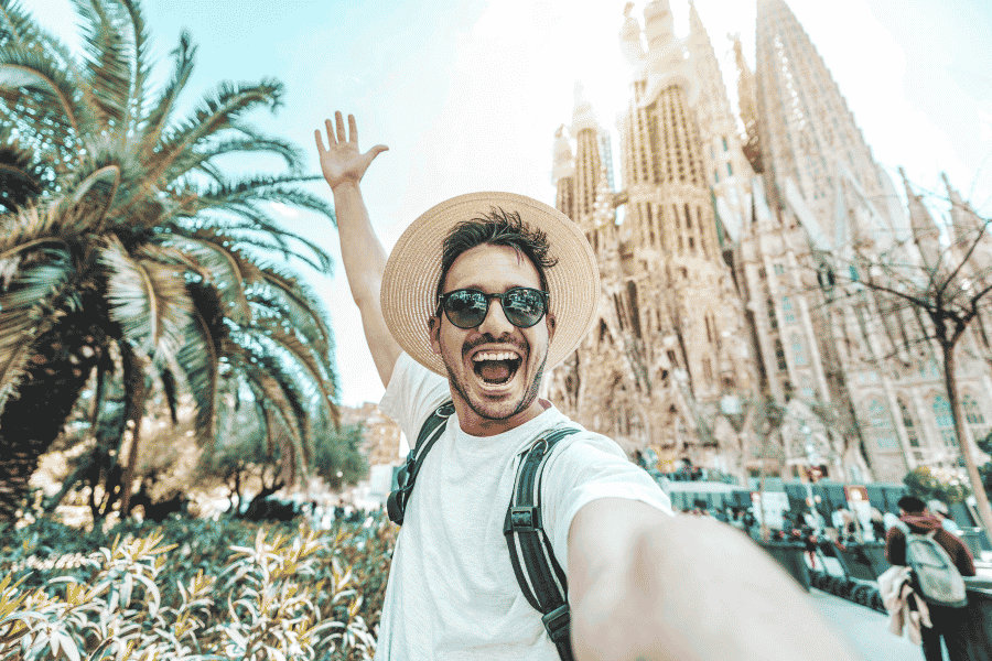Un bărbat entuziasmat cu pălărie și ochelari de soare își face un selfie ridicând o mână, având în spate palmieri și catedrala Sagrada Família sub un cer senin.