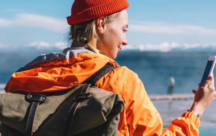 O femeie cu rucsac și căciulă portocalie privește ecranul unui smartphone ținut în mână, în timp ce se află în aer liber, cu un peisaj montan și acvatic în fundal.