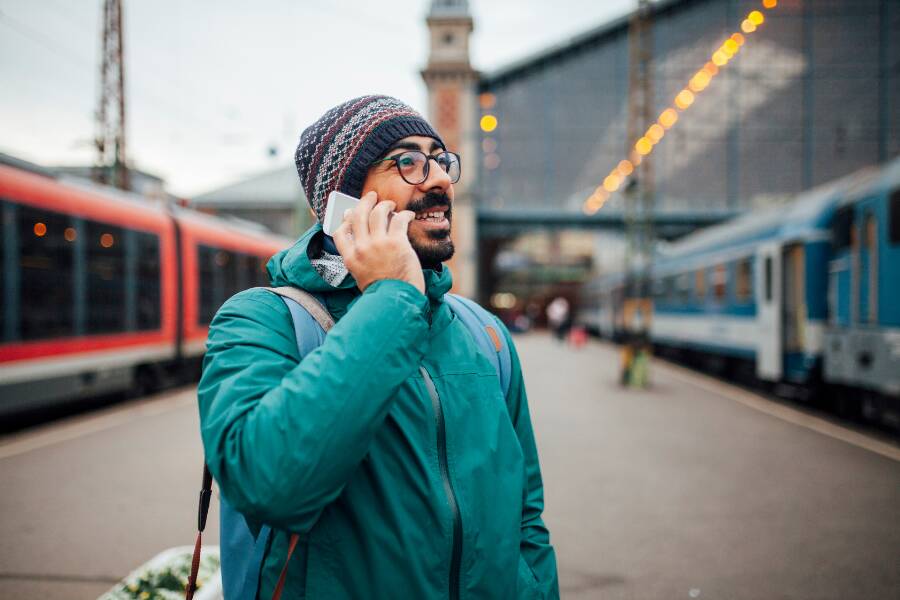 Un bărbat cu ochelari și căciulă vorbește la smartphone pe peronul unei gări, înconjurat de trenuri staționate pe șine în ambele părți și având o clădire în fundal.