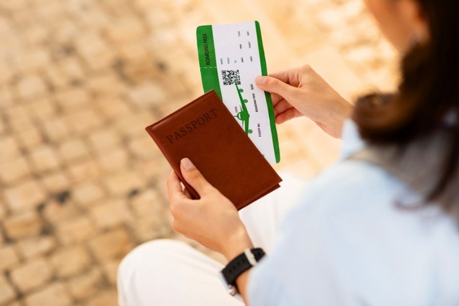 O femeie ține în mâini un pașaport maro pe care scrie PASSPORT și un bilet de avion verde cu alb pe care scrie BOARDING PASS.