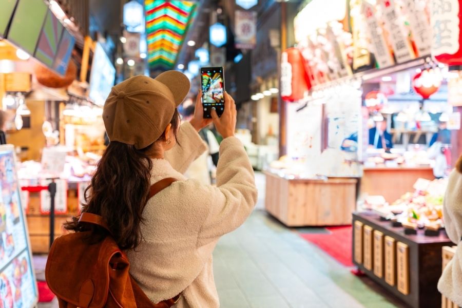 O femeie cu șapcă maro face o fotografie cu telefonul mobil într-o piață acoperită plină de tarabe tradiționale, felinare colorate și lumini vii.