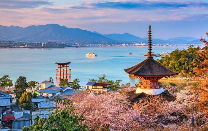 O pagodă japoneză tradițională și o poartă torii se înalță lângă apă, înconjurate de cireși înfloriți, în timp ce un feribot traversează golful pe fundalul munților.