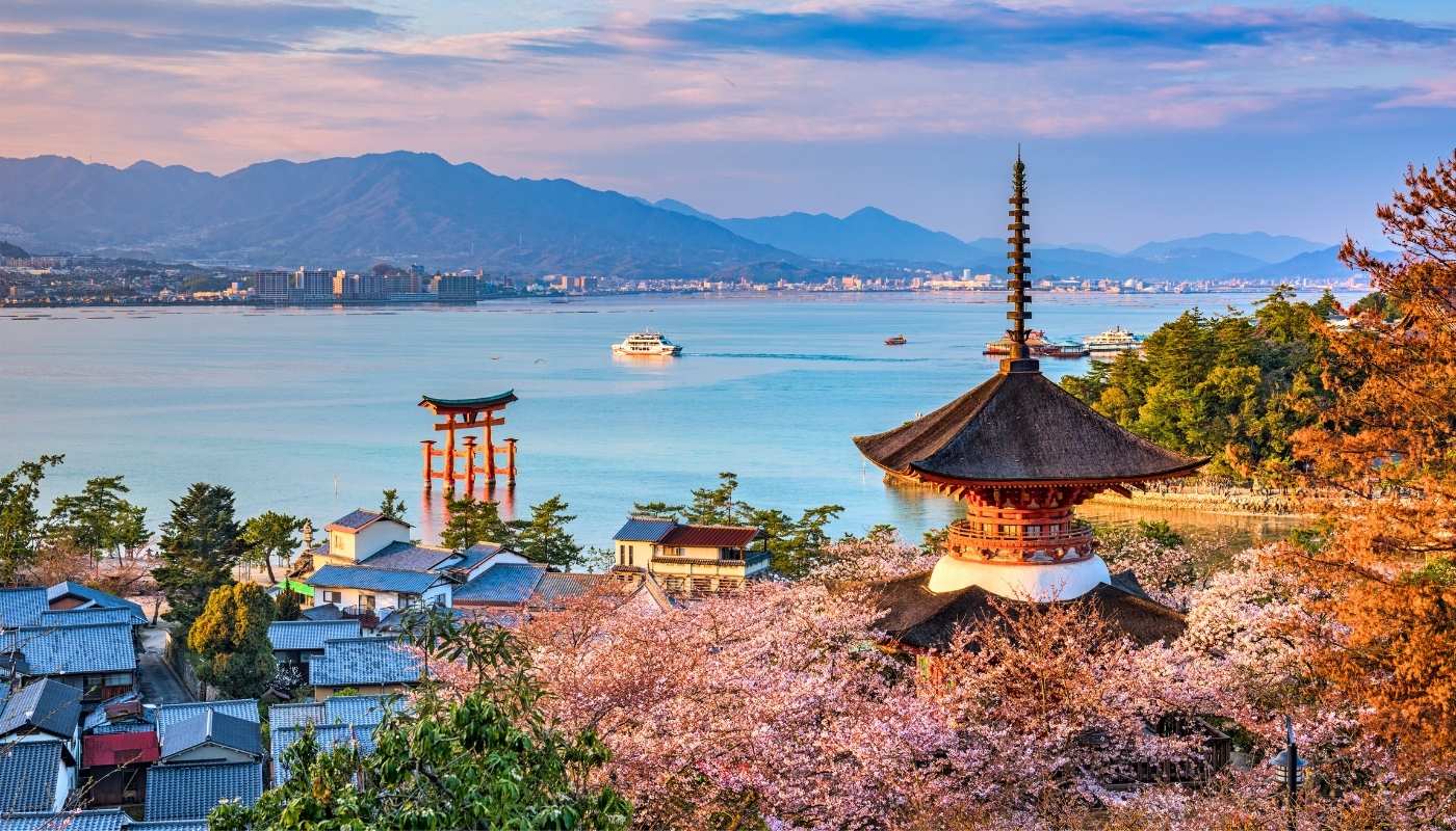 O pagodă japoneză tradițională și o poartă torii se înalță lângă apă, înconjurate de cireși înfloriți, în timp ce un feribot traversează golful pe fundalul munților.